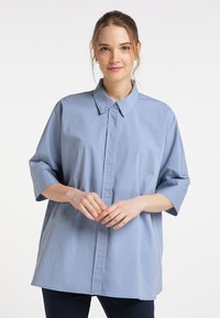 Vaaleansininen ylipitkä paita, jossa on terävä kaulus, napitettava etuosa ja kolme neljännesosaa käärittyjä hihoja. Sujuva kangas, jossa on minimaalinen rakenne.