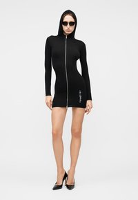 Γυναίκα που φοράει εφαρμοστό μαύρο μίνι φόρεμα με κουκούλα και μπροστινό φερμουάρ, γυαλιά ηλίου και μαύρα ψηλοτάκουνα, στεκόμενη μπροστά σε απλό φόντο.