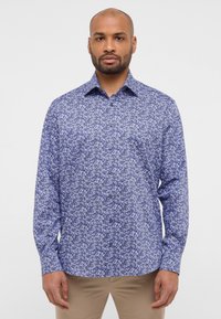 Langearm shirt van blauwe stof met een witte bloemmotief, met een button-down kraag en zwarte knopen, gecombineerd met beige broeken.