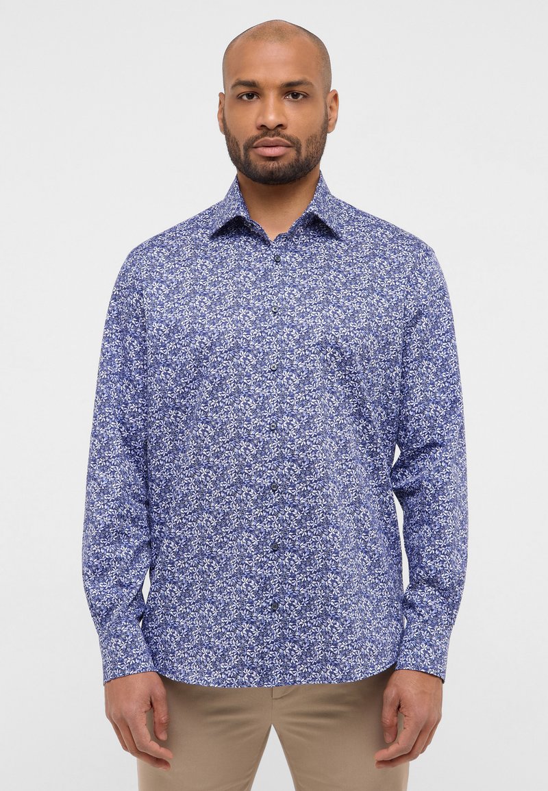 Langearm shirt van blauwe stof met een witte bloemmotief, met een button-down kraag en zwarte knopen, gecombineerd met beige broeken.