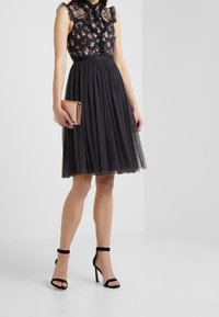Robe noire à motifs floraux avec un décolleté volanté et un empiècement transparent, jupe en tulle plissé, accompagnée d'une pochette métallique et de sandales à talons noirs.