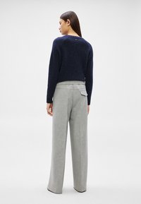 Sieviete, kas stāv ar muguru, valkā tumši zilu džemperi un gaiši pelēkas platām kājām bikses ar aizmugurējo kabatas vāciņu, stāvot pret vienkāršu balto fonu.