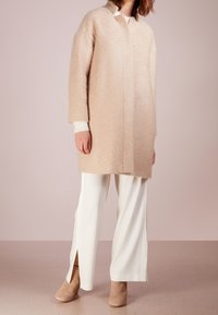 Cappotto oversize beige peloso con colletto, maniche lunghe e dettaglio delle cuciture verticali, abbinato a pantaloni bianchi e stivaletti beige con tacco.