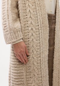 Cardigan beige lavorato a maglia con motivi a cavo texturizzati, dotato di maniche lunghe e vestibilità rilassata, indossato sopra una maglietta chiara e pantaloni a fantasia.