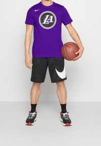 T-shirt Nike violet avec le logo "LA", shorts noirs avec le swoosh blanc de Nike, chaussures de basketball noires et rouges, et ballon de basket tenu dans la main droite.