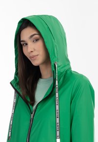 Giacca impermeabile verde con cappuccio, dotata di chiusura con zip e cordini a contrasto etichettati "LOVE". Texture liscia, design leggero.