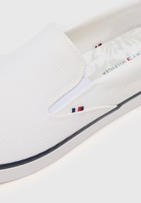 White slip-on shoes with a textured canvas upper, featuring a small logo in navy, red, and white on the side and a black rubber sole.