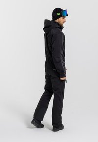 Czarny kombinezon narciarski z kapturem, elastycznymi mankietami i zapinanymi na zamek kieszeniami. W zestawie czarna czapka beanie i lustrzane gogle narciarskie.