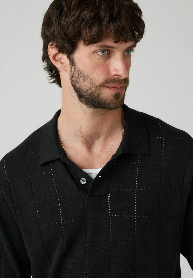 Schwarzes gestricktes Polohemd mit quadratischem Muster und Knopfkragen. Es zeigt ein strukturiertes Finish mit einem subtilen Glanz.