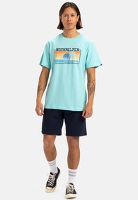 Camiseta de algodón turquesa con un estampado gráfico que incluye un atardecer, una palmera y olas. Combinada con pantalones cortos navy y zapatillas negras.
