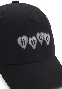 Černá látková čepice s vykrojeným kšiltem, na přední straně stříbrný vyšívaný motiv srdcí a nápis "HUGO".