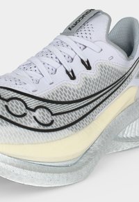 Λευκό και γκρι αθλητικό παπούτσι με μαύρο λογότυπο, λευκά κορδόνια, ανθεκτικό επάνω μέρος από διχτυωτό υλικό και μαλακή σόλα σχεδιασμένη για τρέξιμο.