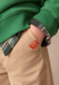 Zelená mikina so stehennými manžetami vrstvená cez kockovanú košeľu, skombinovaná s béžovými nohavicami s oranžovou nášivkou na vrecku.