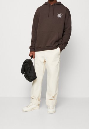 Felpa marrone con cappuccio e grafica del sole, tasca frontale a sacco, materiale in cotone. Abbinata a pantaloni crema e sneakers bianche, con una borsa nera in mano.