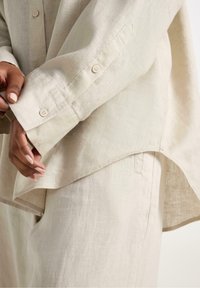 Beige linned skjorte med opprullet ærme, der har en buet kant. Stoffet har en subtil tekstur, og knapperne er synlige på ærmet.