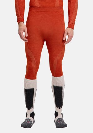 Homme portant des leggings thermiques orange et des chaussettes de ski hautes blanches avec des motifs noirs et gris, debout devant un fond uni.