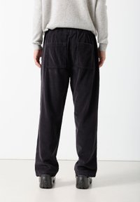 Osoba odjevena u široke crne kord hlače i svijetlo sivu sweater, stojeći bosonoga na jednostavnoj bijeloj pozadini.