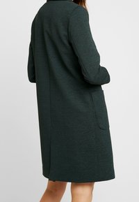 Mørkegrøn frakke med tekstureret stof, lange ærmer og en enkelt frontlomme. Lige snit, knælængde design med klassisk krave.