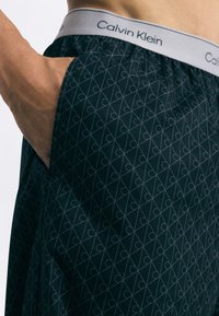 Čierne boxerky Calvin Klein s geometrickým vzorom, s šedým pásom, na ktorom je opakovaný názov značky. Mäkká textúra látky.
