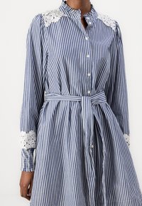 Blå och vit randig klänning med långa ärmar, spetsdetaljer på axlar och handleder, knäppning fram och midja med bälte för form.