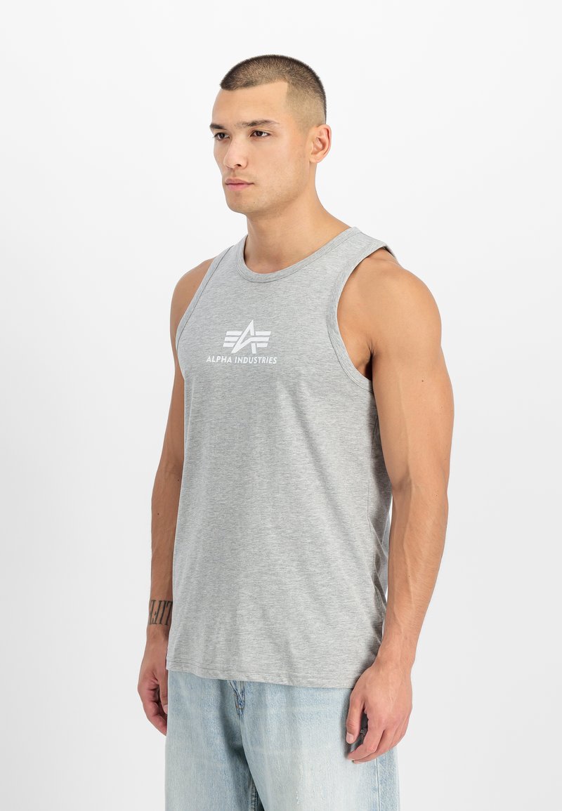 Bărbat cu păr scurt purtând un maiou gri fără mâneci Alpha Industries și blugi bleu deschis, stând în picioare pe un fundal alb.