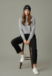 Young woman wearing a black beanie, gray sweater, black pants, and white sneakers, sitting on a wooden stool against a gray background.