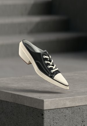 Sort og hvid lavhælet sko med snørebånd, svævende over en grå betonflade med trapper i baggrunden.