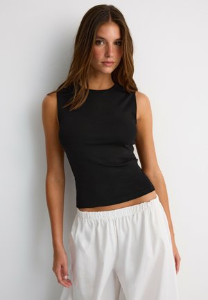 Jeune femme aux cheveux bruns raides portant un haut noir sans manches et un pantalon blanc ample, debout contre un mur blanc.