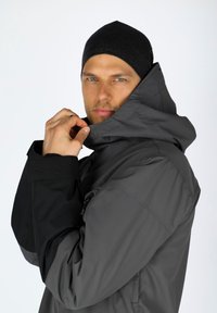 Grijze waterdichte jas met een hoge kraag en zwarte accenten op de mouwen; heeft een gladde textuur en ritssluitingen, gedragen met een zwarte beanie.