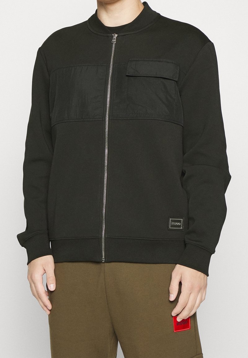 Homme portant une veste noire zippée avec une poche poitrine et un pantalon vert olive avec un patch rouge sur la cuisse, debout devant un fond uni.