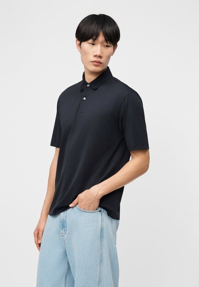 Νεαρός άνδρας που φοράει μαύρο πόλο με κοντά μανίκια και ανοιχτόχρωμο μπλε τζιν, στέκεται με το ένα χέρι στην τσέπη, κοιτάζοντας πλάγια.