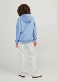 Light blue hooded sweatshirt with a textured finish, paired with light denim jeans. View from the back, emphasizing the relaxed fit.