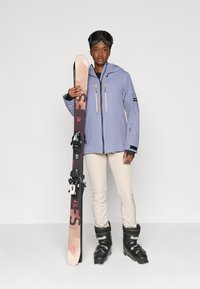 Skijack in lichtblauw met twee gele ritszakken; crèmekleurige broek; zwarte skischoenen; houdt skies vast met een gradient ontwerp.
