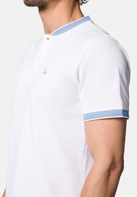 Biała koszulka polo z krótkim rękawem, z niebieskim kołnierzykiem w jodełkę oraz wykończeniem rękawów, z zapięciem na guziki przy szyi i subtelnym logo na piersi.