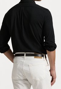 Černá košile s dlouhým rukávem a klasickým límečkem; ve spojení s bílými džínami z denimu. Košile má hladkou texturu a džíny mají zadní kapsy.