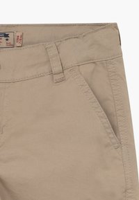 Pantalons en coton couleur tan avec poche frontale visible, passant de ceinture et coutures zippées. Petite étiquette de taille en tissu partiellement visible à l'intérieur de la ceinture.