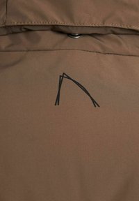 Tejido de chaqueta marrón con una textura suave, que presenta un diseño de logo abstracto negro en la zona del pecho y un detalle de cierre a presión.