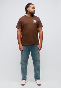 Άνδρας με κοτσίδες, φορώντας καφέ μπλουζάκι με λευκό λογότυπο, μπλε τζιν και λευκά αθλητικά παπούτσια, στέκεται και κοιτάζει προς το πλάι.