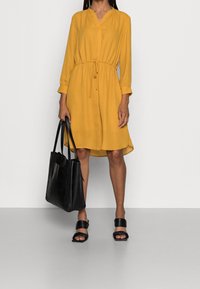 Vestido mostaza de manga larga, con cuello abotonado, cintura ceñida y patrón de lunares. Bolso de mano negro y sandalias de slide negras.