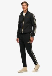 Man in een zwarte trainingspak met gouden strepen, beige hoge sneakers en een horloge, staande tegen een witte achtergrond.