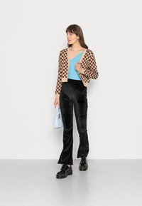 Cardigã xadrez castanho sobre uma blusa azul, combinado com calças flared de veludo preto e sapatos de plataforma, segurando uma mala azul clara.