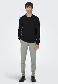 Camisa polo negra de manga larga con cuello y tapeta de tres botones, combinada con pantalones grises claros ajustados y botas negras de tobillo.