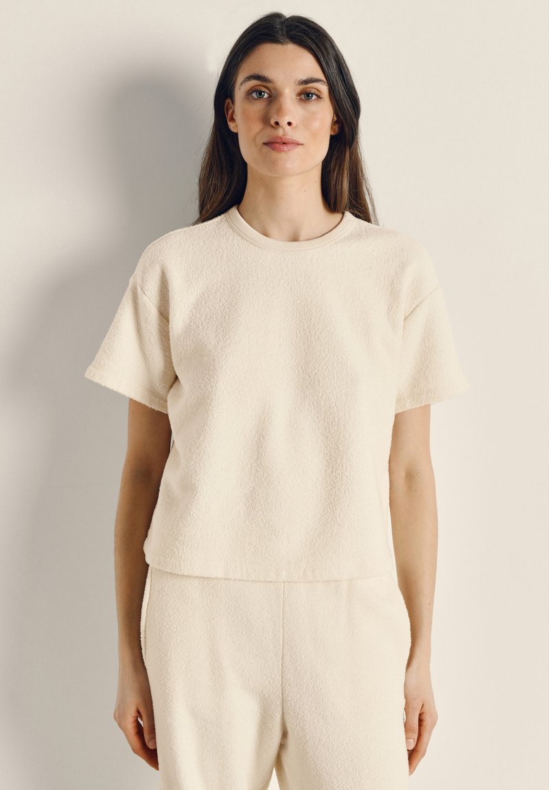 Lysebeige kortærmet top i tekstureret stof med rund halsudskæring. Den har en afslappet pasform og er kombineret med matchende bukser.