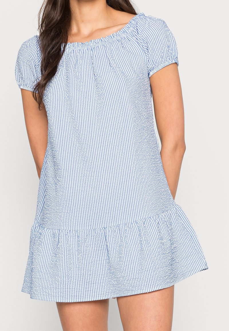 Femme portant une robe à manches courtes, épaules dénudées, rayée verticalement bleu clair et blanc avec un ourlet à volants, debout devant un fond uni.