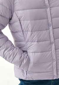 Lyse lavendel pufferjakke med quiltet design, trykknapper og en sidelomme, der har en glat, blank overflade.