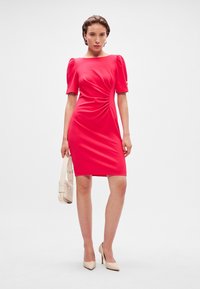 Ružové priliehavé šaty s krátkymi volánovými rukávmi, detailom na bokoch, dĺžka po kolená, v kombinácii s béžovými vysokými podpätkami a malou kabelkou.