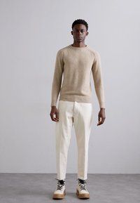 Beige kötött pulóver bordás mandzsettával, fehér tapered nadrággal és fekete fűzőjű bézs csizmával párosítva. Simább tapintás, modern szabás.