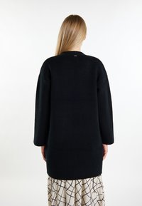 Schwarzer übergroßer Pullover mit Rundhalsausschnitt, langen Ärmeln und weicher Textur. Hat ein einfaches Design und wird über einem gemusterten Rock getragen.