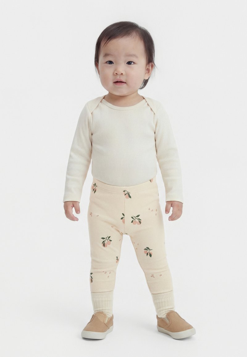 Νήπιο όρθιο, φοράει μπεζ μακρυμάνικη μπλούζα, μπεζ κολάν με ροδακινί ανθισμένο σχέδιο, μπεζ κάλτσες και καφέ παπούτσια χωρίς κορδόνια.