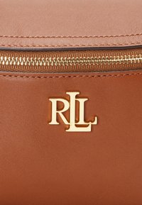 Smeđi kožni dodatak sa zlatnim patentnim zatvaračem i zlatnim RL logotipom centriranim ispod patentnog zatvarača.